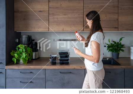 Young woman with healthy diet plan for weight loss, delivery of daily ready meal menu. Woman using meal tracker app on phone. Balanced portion with dish. Pre-cooking. Young woman with healthy diet plan for weight loss, delivery of daily ready meal menu. Woman using meal tracker app on phone. Balanced portion with dish. Pre-cooking. 121968493