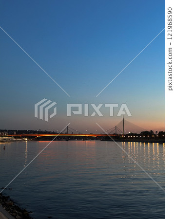 Twilight view of a lit bridge over the river in Belgrade, showcasing serene reflections on the calm water. Harmony of urban architecture in Serbia and nature in the evening fading. Danube river 121968590
