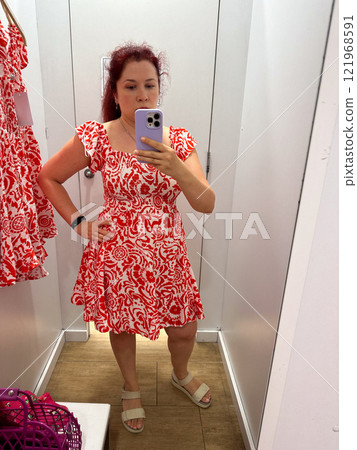 Millennial woman trying on a vibrant summer dress in a fitting room, capturing her reflection in the mirror. Experience of personal shopping and fashion choices in a casual retail setting 121968591