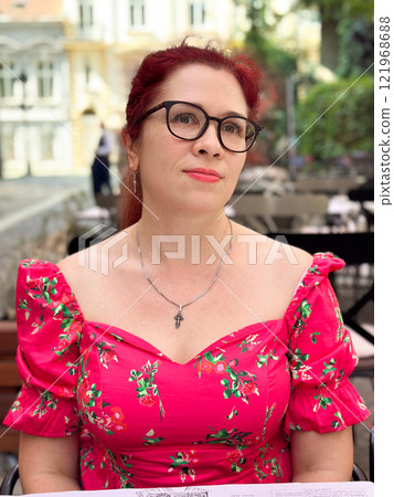 Portrait confident woman in a bright pink floral dress sits outdoors, looking with a calm yet poised expression. Millennial female and summer lifestyle Portrait confident woman in a bright pink floral dress sits outdoors, looking with a calm yet poised expression. Millennial female and summer lifestyle 121968688