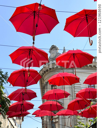 Red umbrellas suspended against a clear blue sky, whimsical and artistic atmosphere. Fusion of creativity and urban architecture, design exterior of outdoor restaurant and hotel. 121968698