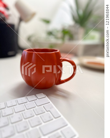 Cozy autumn workspace with a vibrant pumpkin shaped mug on a clean desk. Minimalist decor and warm tones create a relaxing atmosphere, perfect for work or leisure during the fall season 121968844