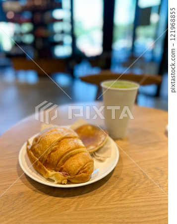 Cozy breakfast scene with a croissant, pancake and matcha latte served on a wooden table. Blurred background of a cafe adds depth and evokes a sense of warmth and relaxation in a casual dining setting 121968855
