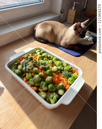 Cozy kitchen moment with a siamese cat basking in sunlight next to a colorful tray of fresh vegetables. Harmonious blend of food preparation and peaceful companionship in a warm domestic setting 121968879