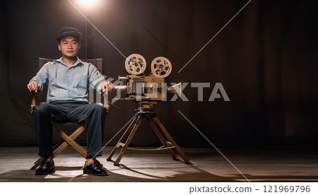 A man sits in a chair in front of a movie camera 121969796