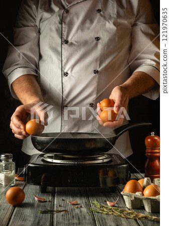 Hands expertly hold fresh eggs above a hot skillet, ready for cooking. The ambiance is warm and inviting, with kitchen tools and ingredients arranged nearby, enhancing the culinary atmosphere 121970635