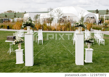 Wedding ceremony setup featuring white columns, flower arrangements, and chairs on a green lawn 121973972