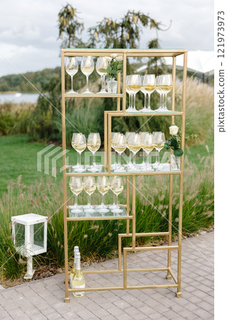 Golden metal shelves displaying glasses of white wine and bottles for an outdoor event Golden metal shelves displaying glasses of white wine and bottles for an outdoor event 121973973