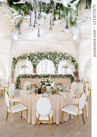 Elegant wedding reception hall featuring round tables, white and gold chairs, and floral arrangements Elegant wedding reception hall featuring round tables, white and gold chairs, and floral arrangements 121973975