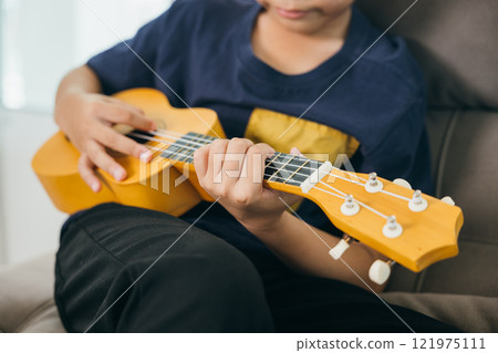 Fingers glide over the strings of a yellow ukulele in this close-up. The young musician sits on a couch, blending creativity and relaxation in a cheerful family setting. 121975111
