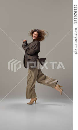 Dynamic shot of young attractive lady, with brunette curly hair,dancing of joy, dressed formalwear against grey studio background, Dynamic shot of young attractive lady, with brunette curly hair,dancing of joy, dressed formalwear against grey studio background, 121976572