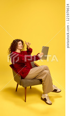 Cheerful smiling young woman sitting in chair and taking selfie on tablet gesturing peace sign against yellow studio background. Cheerful smiling young woman sitting in chair and taking selfie on tablet gesturing peace sign against yellow studio background. 121976614