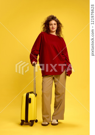 Confident young woman dressed casual, oversized clothes posing holding yellow suitcase against sunny studio background. Confident young woman dressed casual, oversized clothes posing holding yellow suitcase against sunny studio background. 121976628