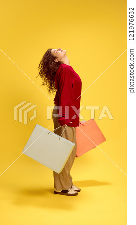 Joyful young fashionista, stylish woman holding purchases and posing, dressed casually, against yellow studio background. 121976632