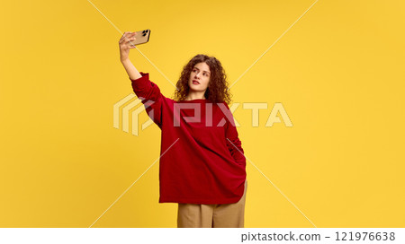 Casually dressed, young brunette woman with curly hair, taking selfie against sunny yellow studio background, Self-love. 121976638