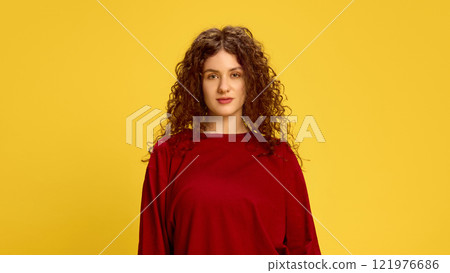 Calm and serious, young woman, with curly brunette hair, dressed red t-shirt, looking at camera against vibrant yellow background. Headshot. Calm and serious, young woman, with curly brunette hair, dressed red t-shirt, looking at camera against vibrant yellow background. Headshot. 121976686
