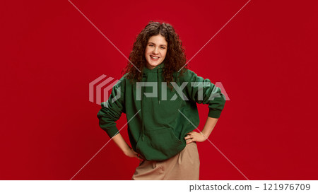 Cheerful smiling young woman with curly hair posing, dressed casual stylish clothes, holding hands on hips against bold red background. 121976709