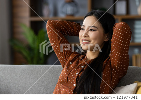 Asian woman in polka dot blouse enjoying relaxation at home, smiling as she leans back on sofa. Her peaceful expression conveys happiness and contentment in cozy setting. 121977341