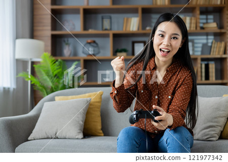 Asian woman playing video games at home on couch, holding controller with excitement. Joyful gaming experience, expressing thrill and competitive spirit in comfortable living room setting. 121977342