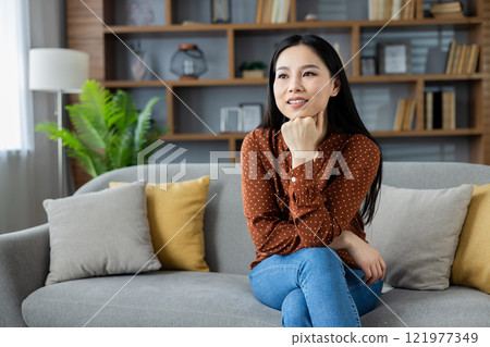 Asian woman relaxing at home, sitting on sofa with thoughtful expression. Surrounded by books, cushions, and plants, conveying calm and contemplation in a stylish living room setting. Asian woman relaxing at home, sitting on sofa with thoughtful expression. Surrounded by books, cushions, and plants, conveying calm and contemplation in a stylish living room setting. 121977349