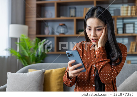 Asian woman experiencing shock while reading phone message in home setting. Seated on sofa, she appears worried and surprised. Modern living room provides calm yet serious atmosphere. 121977385