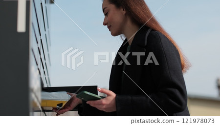 Woman receiving parcel from post terminal machine using smartphone outdoors. Parcel delivery machine. Mail delivery and post service, online shopping, e commerce concept. 121978073