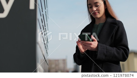 Woman receiving parcel from post terminal machine using smartphone outdoors. Parcel delivery machine. Mail delivery and post service, online shopping, e commerce concept. 121978074