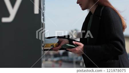 Woman receiving parcel from post terminal machine using smartphone outdoors. Parcel delivery machine. Mail delivery and post service, online shopping, e commerce concept. 121978075