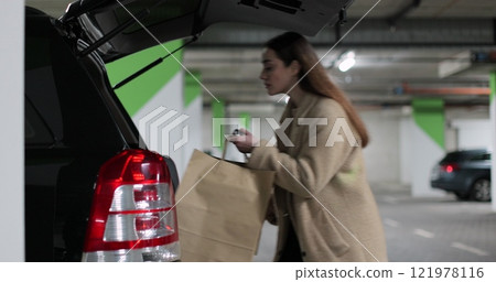 Beautiful woman shopper walks through mall parking lot holds cardboard bag in hand open trunk of car. Beautiful woman shopper walks through mall parking lot holds cardboard bag in hand open trunk of car. 121978116