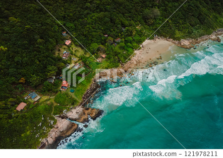 Drone view of a secluded beach with turquoise waters, surrounded by lush forest, rocky coastline, and scattered houses in Brazil. 121978211