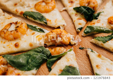 Close-up of seafood pizza slice with shrimp, spinach, and melted cheese on a wooden platter. Close-up of seafood pizza slice with shrimp, spinach, and melted cheese on a wooden platter. 121978843
