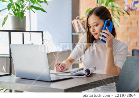 Young woman serious sitting at table with laptop talking on mobile phone in coworking Young woman serious sitting at table with laptop talking on mobile phone in coworking 121978864