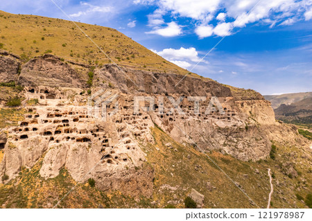 Aerial top view of Vardzia city on sunny summer day. Ancient cave monastery in Erusheti Mountain rocks at Kura River. Monument of medieval Georgian architecture, carved in rough rock. Georgia, Europe. Aerial top view of Vardzia city on sunny summer day. Ancient cave monastery in Erusheti Mountain rocks at Kura River. Monument of medieval Georgian architecture, carved in rough rock. Georgia, Europe. 121978987