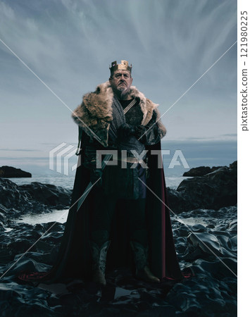 Art collage. Medieval king in golden crown, fur-lined cloak, and armor, holding sword, standing on rocky coastline under dramatic sky 121980225