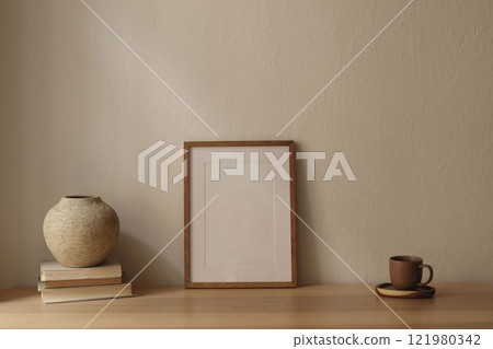 Elegant minimal home office still life. Blank wooden picture frame poster mockup on table. Scandi interior decor. Cup of tea, coffee, old books. Textured vase. Beige wall background. Empty copy space. Elegant minimal home office still life. Blank wooden picture frame poster mockup on table. Scandi interior decor. Cup of tea, coffee, old books. Textured vase. Beige wall background. Empty copy space. 121980342