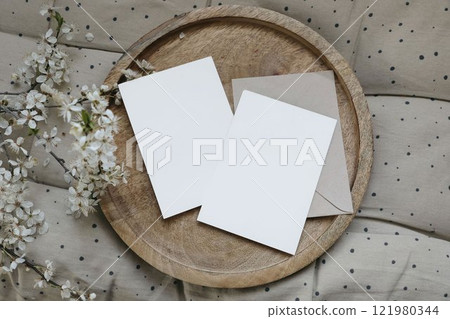 Spring stationery still life. Two empty greeting cards, invitations mockups on wooden tray. Craft envelope. Blurred blossoming cherry plum tree branches. Textile beige background with black dots. Top 121980344
