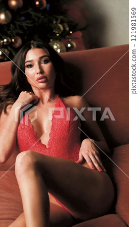 Beautiful brunette posing sexy on camera in red lace bodysuit while sitting on sofa 121981569
