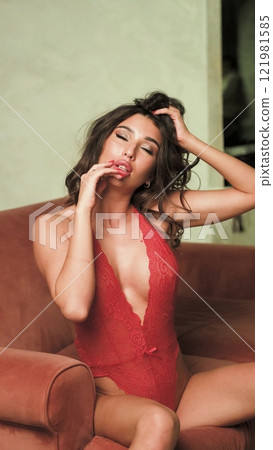 Beautiful brunette posing sexy on camera in red lace bodysuit while sitting on sofa Beautiful brunette posing sexy on camera in red lace bodysuit while sitting on sofa 121981585