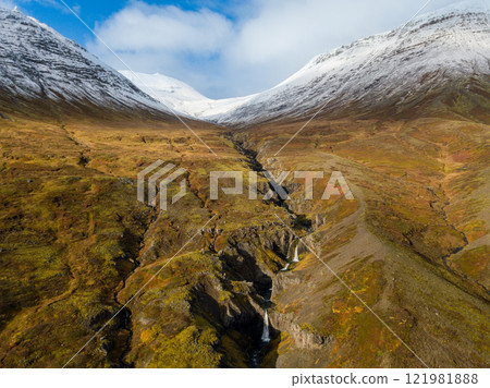 Aerial of Reydarfjordur: Sunlight streaming over a river highlights a vibrant valley leading to majestic snowy mountains in Iceland. Nature's beauty unfolds in a serene landscape filled with color and 121981888
