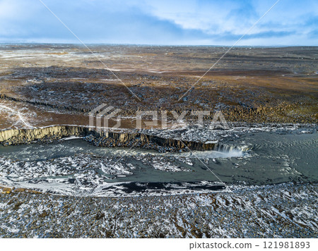 Turquoise glacial river winds through a frozen Icelandic landscape, flowing toward the majestic Dettifoss waterfall, showcasing nature's raw beauty and power from above 121981893