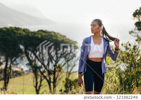 Slim female in fitness attire adjusting her hair while standing in front of stunning view at sunset Slim female in fitness attire adjusting her hair while standing in front of stunning view at sunset 121982249