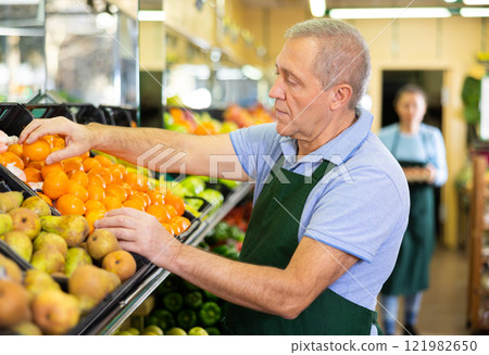 Skillful elderly salesman laying out tangerines in shop 121982650