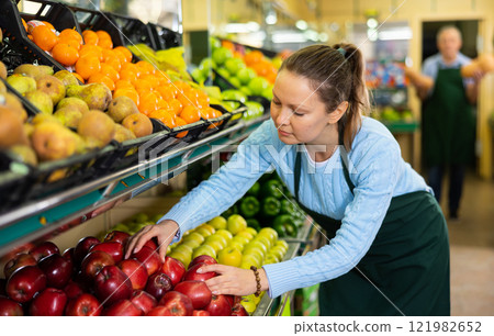 Middle-aged saleswoman putting apples on food stall in grocery store Middle-aged saleswoman putting apples on food stall in grocery store 121982652