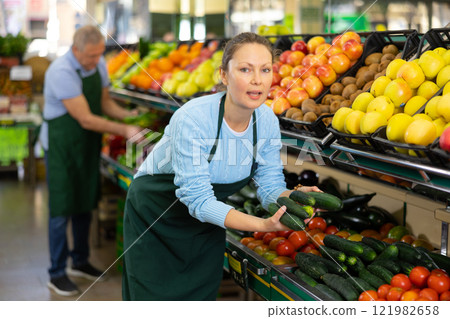 Positive middle-aged woman, skilled market assistant arranging organic cucumber on shelves in hypermarket 121982658