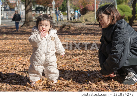 [A one-year-old child stands up, playing in a winter park covered with fallen leaves] 121984272