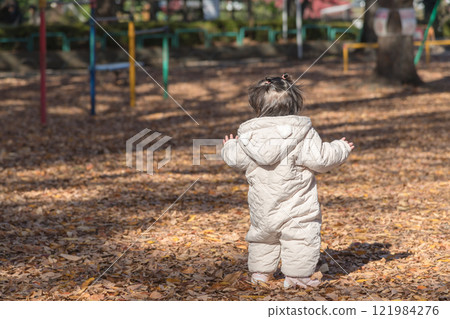 [A one-year-old child playing in a winter park covered with fallen leaves, standing from behind] 121984276