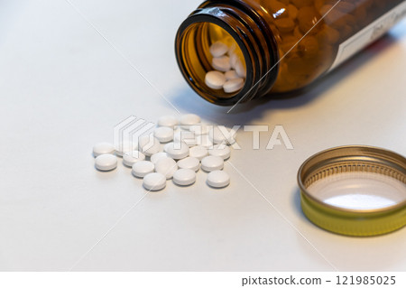 White pills spilled onto a table from an overturned glass bottle 121985025