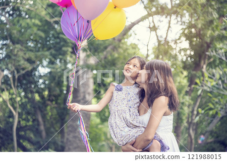 Happiness mother daughter play balloons together in green garden park outdoor lifestyle. Happy family two people enjoy have fun cheerful smile. Mother childhood hand holding colorful balloons outside 121988015