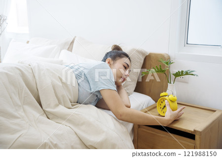 Asian women sleep wake up in white cozy bedroom happy time morning sunrise. Beautiful chinese use hand stop time alarm clock. Happy person lifestyle wake up in morning wellbeing relax sleepy napping 121988150