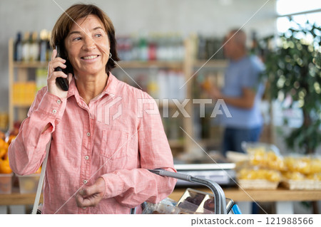 In shop mature woman with market basket in hands talk on phone, call friend. Man buyer in background In shop mature woman with market basket in hands talk on phone, call friend. Man buyer in background 121988566